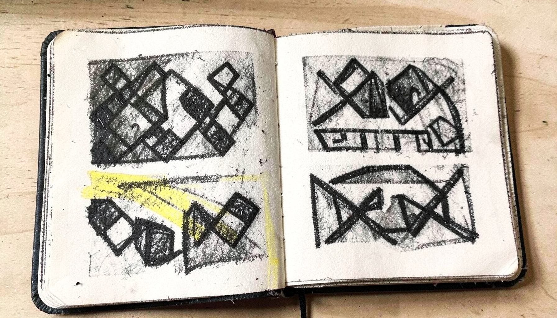 A bold charcoal drawing of a sketchbook open to logo thumbnails, with geometric shapes and letterforms in dark graphite tones against a pale background